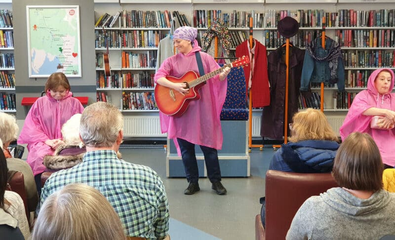 People in a library. A woman stands in a pink waterproof at a microphone, playing a guitar. People sit around listening. Some of them are also in pink waterproofs