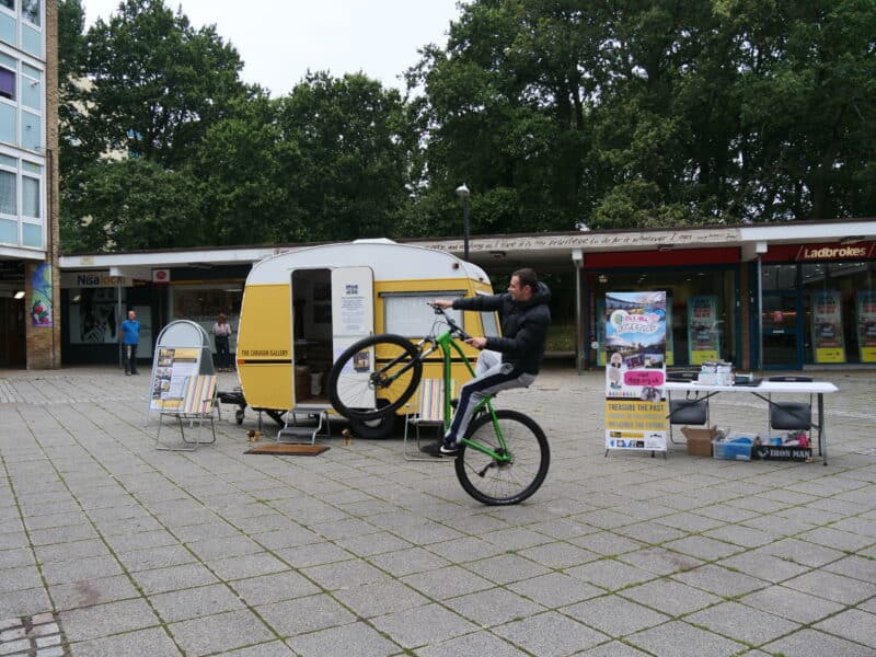 A small yellow caravan sits outside a 1970's shopping precinct. A young man rides a BMX and performs a 'wheelie' in front of the caravan